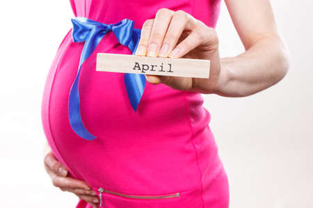 Woman in pregnant wearing pink dress with blue ribbon and showing word april written on wooden cube....