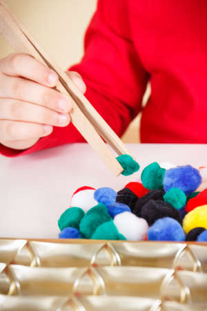 Preschooler playing with small fluffy colorful pompoms and wooden tongs. Development of kids motor...