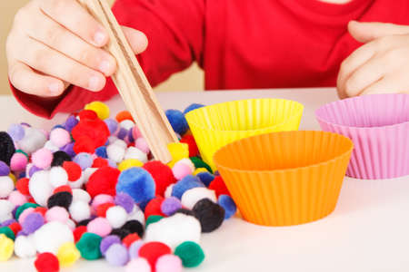 Preschooler playing with small colorful pompoms and wooden tongs. Development of kids motor skills, coordination, creativity and logical thinkingの写真素材