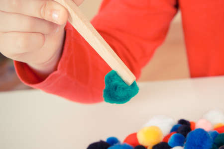 Preschooler playing with small fluffy colorful pompoms and wooden tongs. Development of kids motor skills, coordination, creativity and logical thinkingの写真素材
