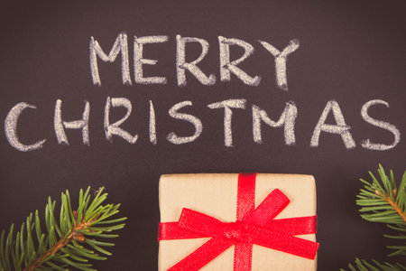 Inscription Merry Christmas, wrapped gift with ribbon and green spruce branches on black background. Decoration for christmas timeの写真素材
