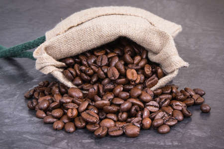 Heap of dark roasted coffee grains in jute bag on concrete structure