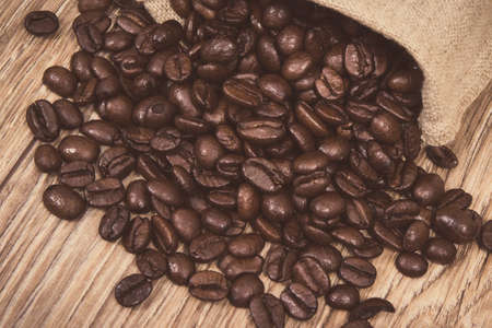 Heap of dark roasted coffee grains in jute bag on wooden background