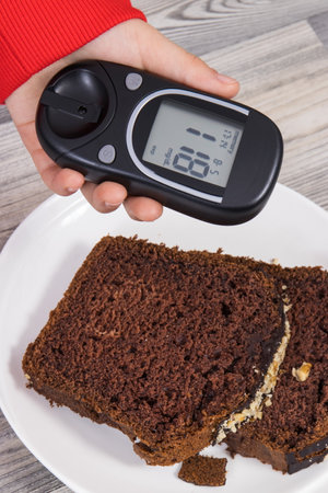 Hand of child holding glucometer with result of measurement sugar level, sweet chocolate cake. Nutrition during diabetes conceptの写真素材