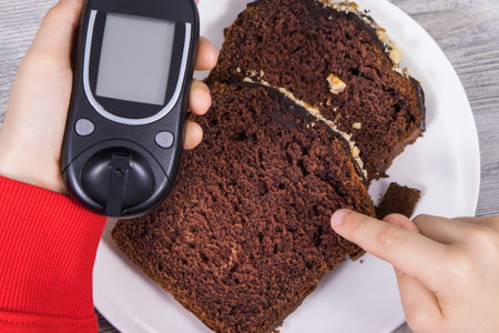 Hand of child holding glucose meter, sweet chocolate cake. Measuring and checking sugar level during diabetesの写真素材