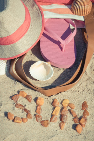 Inscription sun made of amber and different accessories for relax on sand. Straw hat, pink slippers and fluffy towel. Summer time on the beachの写真素材
