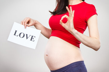 Pregnant woman holding white card with inscription love and showing sign ok. Expecting birth of newborn. Extending family. Motherhoodの写真素材