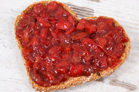 Slice of wholegrain bread with strawberry jam for breakfast. Shape of heart. Old rustic backgroundの写真素材
