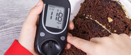 Hand of child holding glucose meter with result of measurement sugar level, sweet chocolate cake. Nutrition during diabetes conceptの写真素材