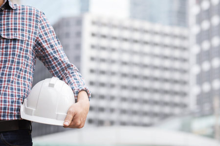 Close up backside view of engineering male construction worker stand holding safety white helmet and wear reflective clothing for the safety of the work operation. outdoor of building background.の写真素材