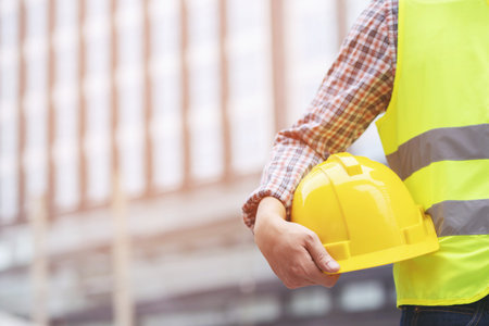 Close up front view of engineering male construction worker stand holding safety white helmet and wear reflective clothing for the safety of the work operation. outdoor of building background.の写真素材