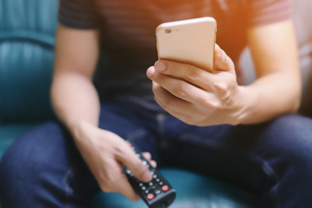 young man using television remote control and mobile smart phone is sitting on a sofa. on or off tv. soft focusの写真素材