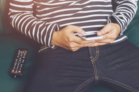 young man using television remote control and mobile smart phone is sitting Search for his favorite channel list on a sofa. on or off tv.の写真素材