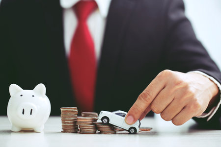 Business man in suit and red necktie holding model of toy car on over a lot of stacked coins - insurance, loan and buying car conceptの写真素材