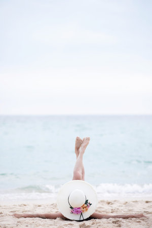 tanning suntanned young woman in bikini wearing straw hats lying on a tropical beach on white sand stretching up slender leg Blue sea in the background.の写真素材