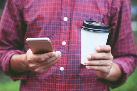 Young male using on cell phone stand in watching message on mobile phone during break hand holding Paper cup of take away drinking coffee hot on the morning in public park.の写真素材