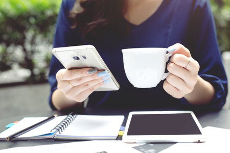 businees woman sitting take a break hand holding cup drink coffee in coffee shop at wooden table, and using smartphone with tablet note book document. Girl browsing internet, chatting blogging.の写真素材