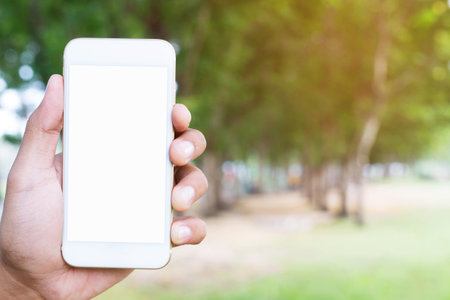 close up people hand holding display white mobile smart phone with blank of empty white screen on outdoor public parks green tree background.の写真素材