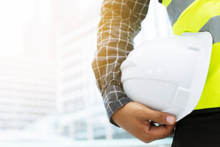 Close up front view of engineering male construction worker holding safety white helmet and wear reflective clothing for the safety of the work operation. outdoor of building background.の写真素材