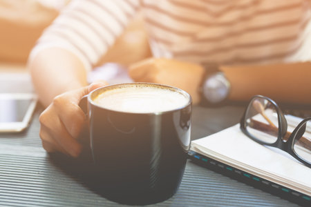 close up people female sit hand holding cup coffee on desk table with book notepad,equipment supplies in the work. copy space. concept to Drink coffee before work to refresh.の写真素材