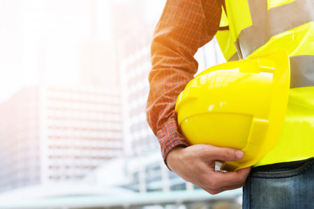 Close up front view of engineering male construction worker stand holding safety yellow helmet and wear reflective clothing for the safety of the work operation. outdoor of building background.の写真素材