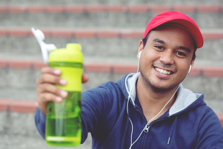 Young man sit listen to music happily break drinking water after running Jogging workout. Thirsty healthy male with fit body drinking water.の写真素材