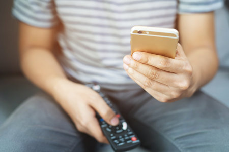 young man using television remote control and mobile smart phone is sitting on a sofa. on or off tv.