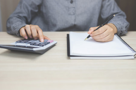 Close up young woman hand is writing in a notebook and using calculator counting making notes...