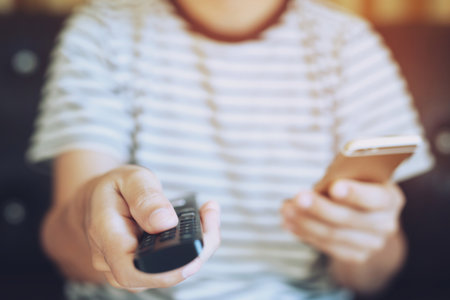 young man using television remote control and mobile smart phone is sitting on a sofa. on or off tv.