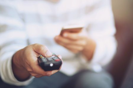 young man using television remote control and mobile smart phone is sitting on a sofa. on or off tv.の写真素材