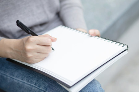 close up hand young woman are sitting on a marble chair. using pen writing Record Lecture note pad into the book in park public.の写真素材
