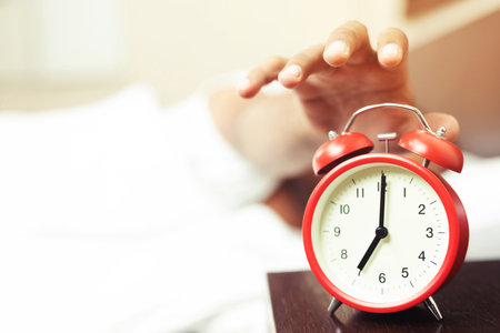 Close up of man lying in bed turning off hand reaching out for alarm clock color red waking up at morning at 7am.の写真素材