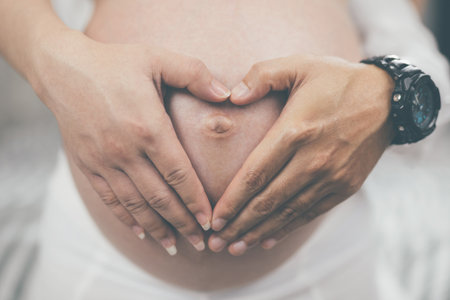 pregnant woman and her husband hand splice hugging the tummy Heart shaped show love for the unborn baby in the belly relaxing at home in bedroom. wait birth date expecting a baby stand in the outdoorsの写真素材