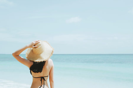 backside young woman skin tan in sunhat and bikini standing with her arms raised to her head...