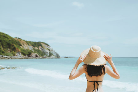 backside young woman skin tan in sunhat and bikini standing with her arms raised to her head enjoying on the beach vacation travel. and enjoy life at sea looking view of beach ocean on hot summer day.の写真素材