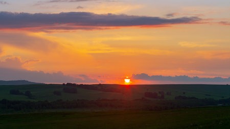 The sun sets on the horizon, the sky and the clouds are illuminated by the orange rays of the sun, summertime, the slopes of the hill are covered with green grass and trees.の写真素材