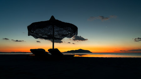 The beginning of the day at the Red Sea. Dark silhouettes of a chaise lounge and an umbrella can be seen on the beach with the orange light from the first rays of the sun and the island of Tiran behind them. Holiday at the resorts of the Sinai.の写真素材