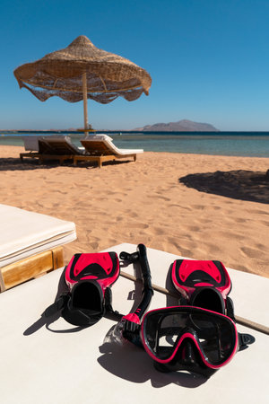 A red mask, snorkel, and fins can be seen in the foreground. A sunny beach with sun loungers and umbrella. Tiran Island. Egypt.の写真素材