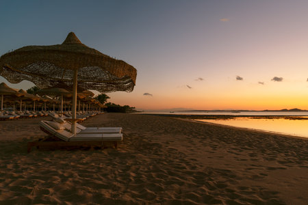 On a resort beach with fine sand, sun loungers and umbrellas sit under an orange sunrise over the sea. High resolution photo.の写真素材