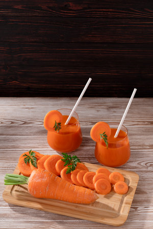 On a wooden table there are carrots, a cutting board with carrot slices, a knife and two glasses of fresh juice with straws. Soft light illuminates the objects.の写真素材