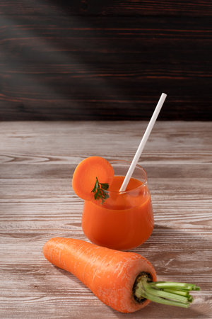 There is a carrot on the wooden table and a glass of fresh juice with a tube. Soft light illuminates the objects.の写真素材