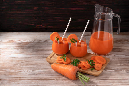 On a wooden table, there are carrots, a jug and two glasses of fresh juice with carrot pieces. Soft light illuminates the objects.の写真素材