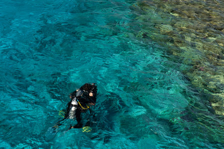 A scuba diver in full gear with an air tank is swimming in clear turquoise water above a coral reef.の写真素材