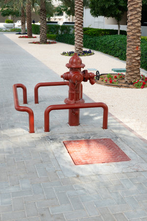 Dubai, UAE - February 4, 2023: Red fire hydrant surrounded by a metal fence on a sidewalk, with palm trees, white gravel, and a green lawn in the background.のeditorial素材