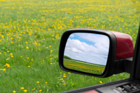 Side view mirror of a red car reflecting a beautiful spring landscape with blue sky and clouds over a green field of yellow dandelions.の写真素材