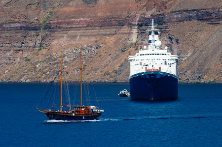 Sailboat sails in front of a cruise ship on the coast of Santorini, Greece. Island cliffs in the backgroundの写真素材