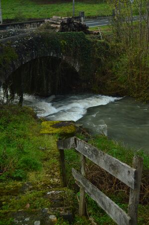 Beautiful Postcard With A Brave River, A Roman Bridge And A Mountain Of Freshly Cut Logs Wet By The...
