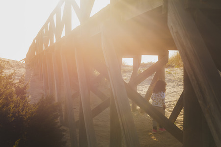 little girl playing between the wooden pillars of a catwalk that leads to the beach with the sunset light creating a beautiful flareの写真素材