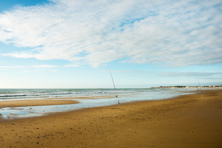two fishing poles nailed in the wet sand on the shore of the beach with the clouds passing through the blue sky and some cliffs in the backgroundの写真素材