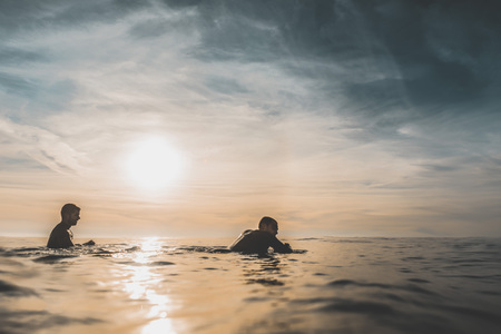 Two surfer friends patiently wait for a wave over the calm sea at sunset. Extreme water sports and...